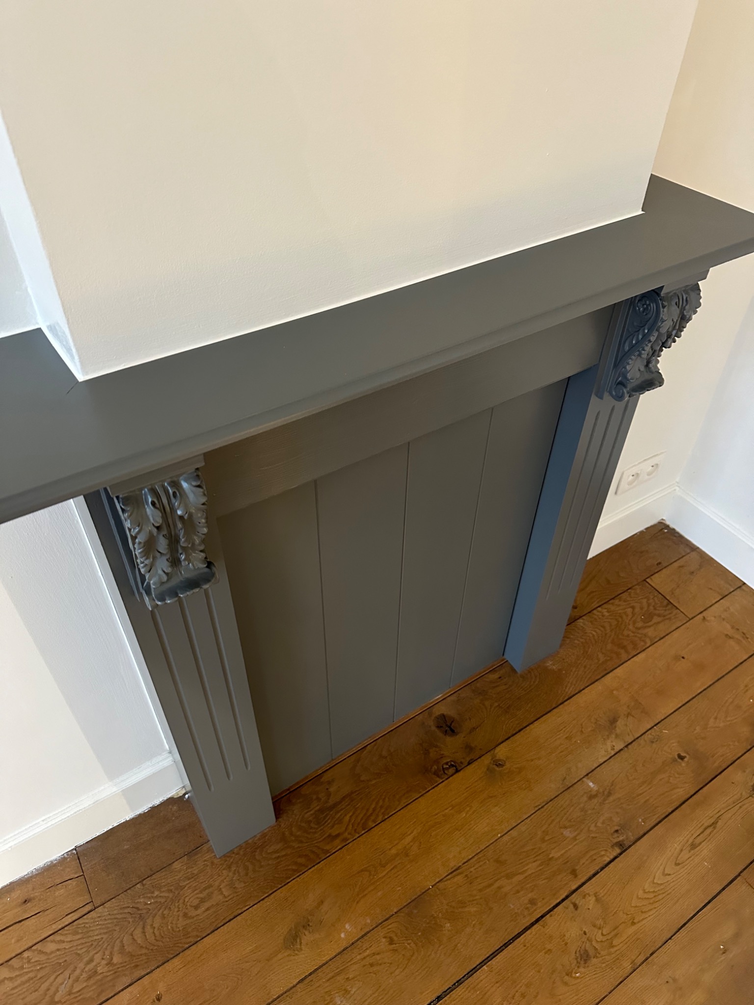 Szary, odnowiony portal kominkowy z dekoracyjnymi elementami na tle białej ściany i drewnianej podłogi.