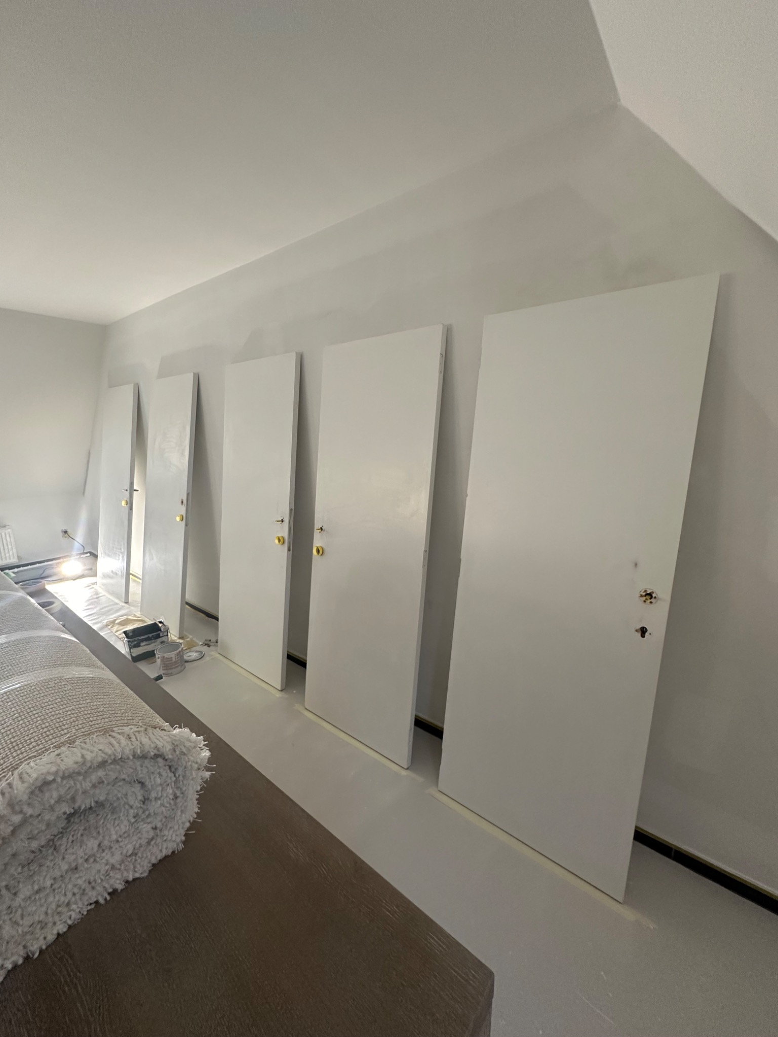 Białe, świeżo pomalowane drzwi z zamontowanymi złotymi klamkami, ustawione w rzędzie w jasnym, przygotowanym do prac pomieszczeniu, widoczne ślady taśmy malarskiej na podłodze i narzędzia malarskie...