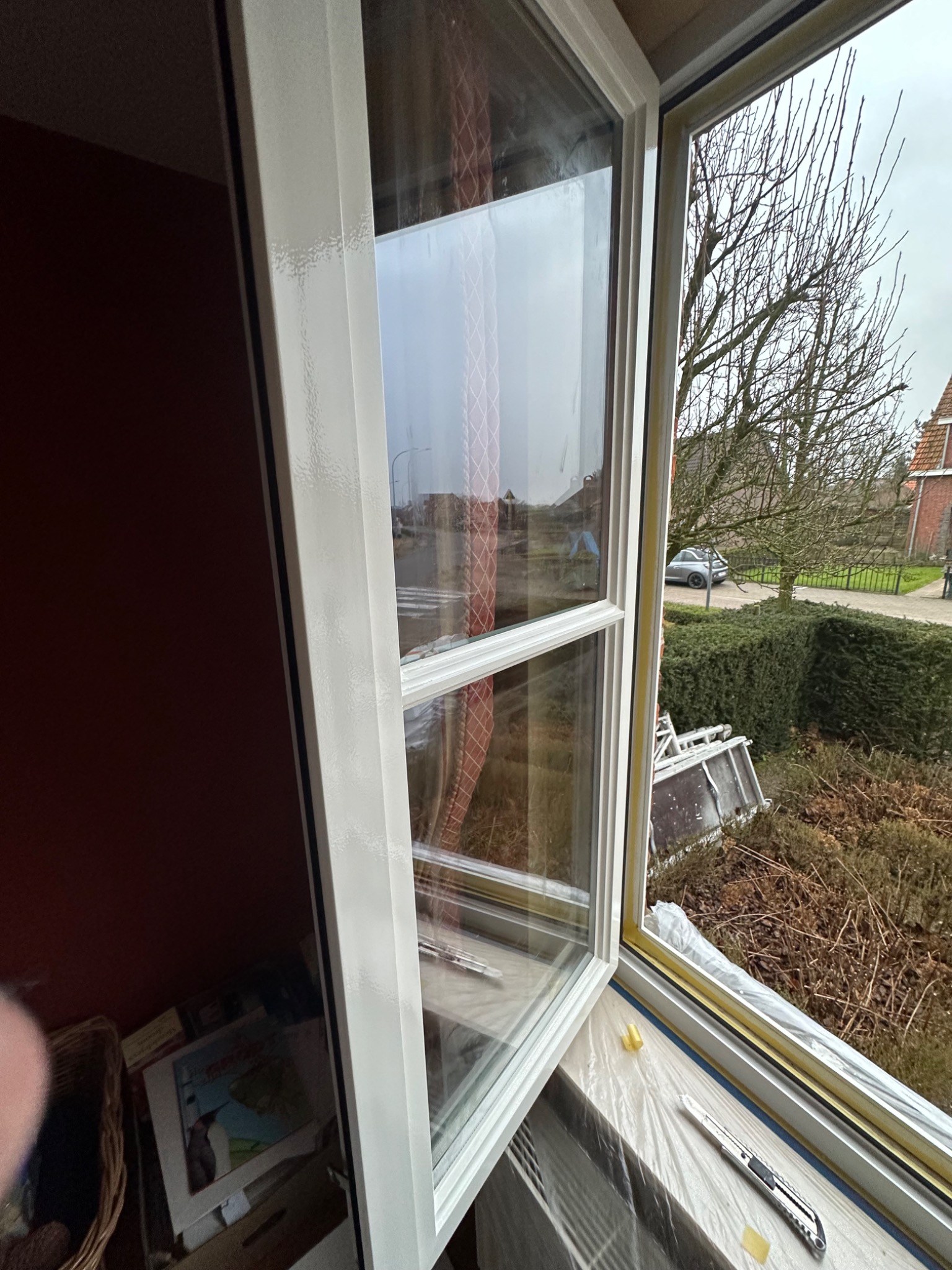 Białe okno PCV, częściowo otwarte, z widoczną uszczelką, folią ochronną i nożykiem na parapecie; za oknem widok na ulicę z drzewami i domami.