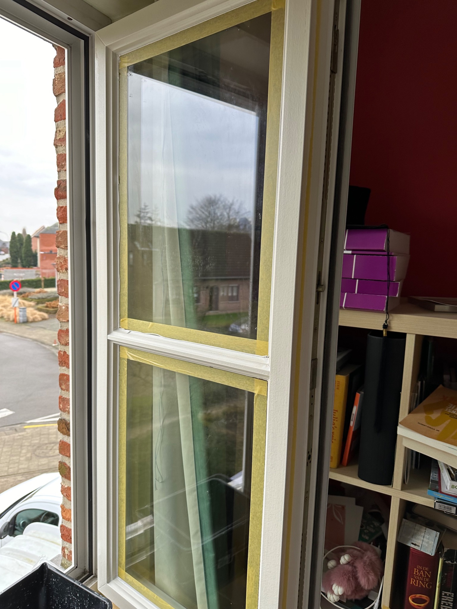 Przygotowane do malowania, dwuskrzydłowe okno z białymi ramami zabezpieczone żółtą taśmą malarską, widok na ulicę z ceglanym budynkiem i częściowo zasłonięty regał z książkami w tle.