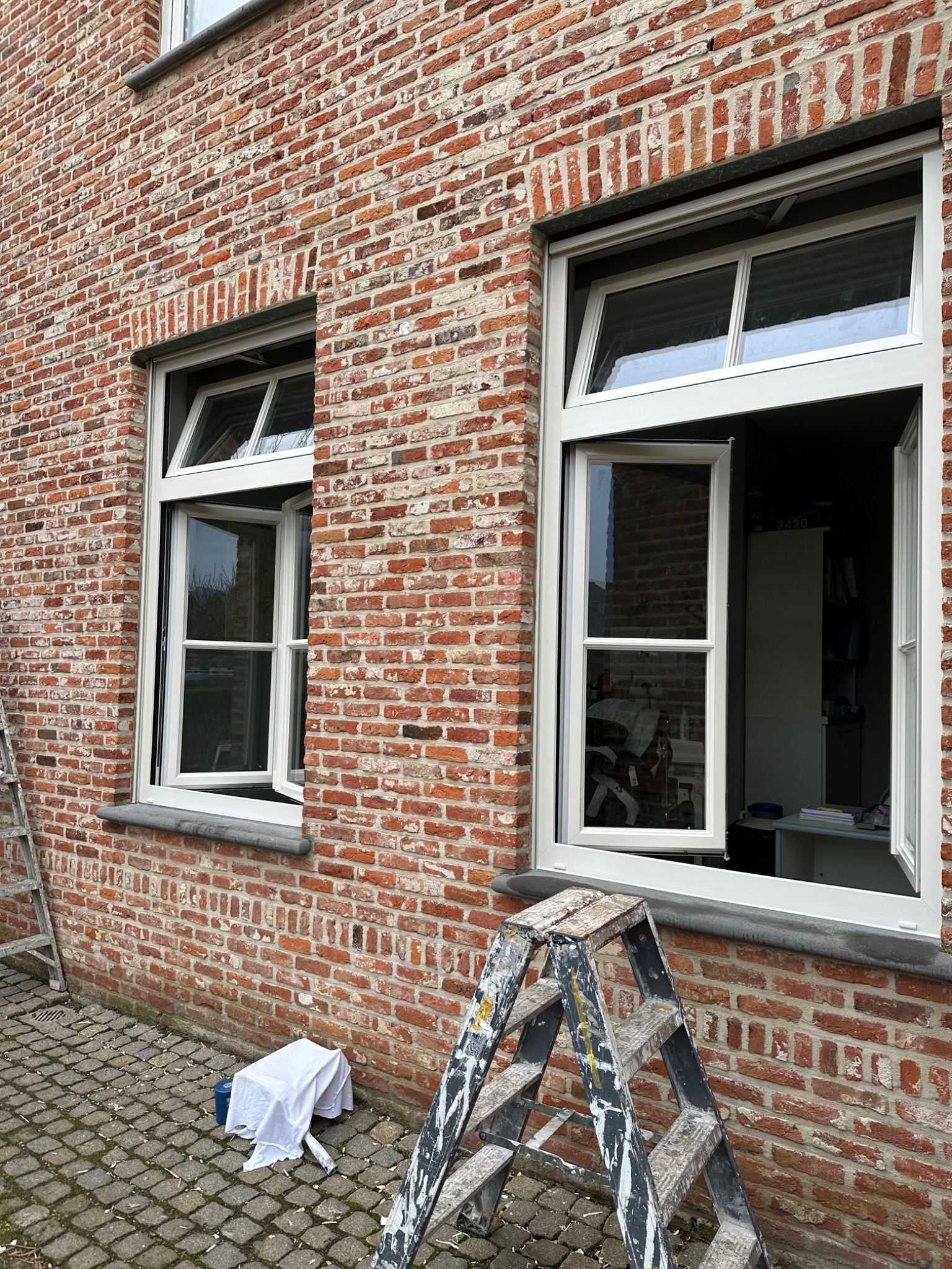 Dwa okna PCV z białymi ramami w murze z czerwonej cegły, jedno okno otwarte, drabina malarska i zabezpieczona plandeką puszka na brukowanym podłożu.