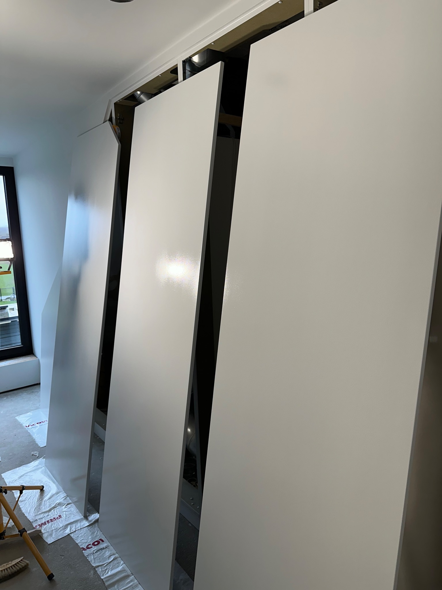 Świeżo pomalowane na biało płyty gipsowo-kartonowe, oświetlone od góry, opierają się o ścianę podczas remontu mieszkania, zabezpieczone folią podłogową.