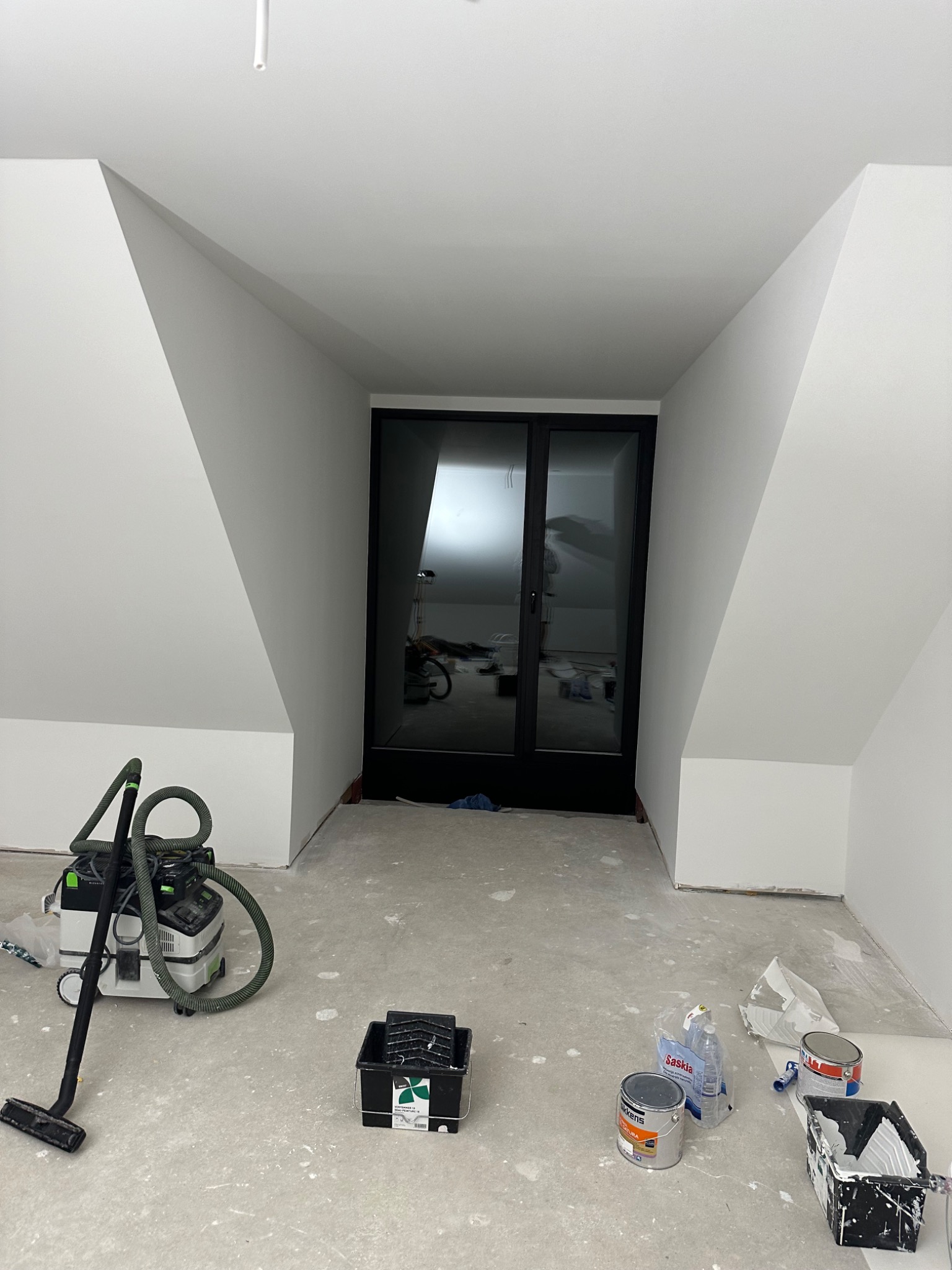 Poddasze po świeżym malowaniu, widoczne białe ściany ze skosami, czarne drzwi przesuwne, odkurzacz przemysłowy, puszki po farbie i kuwety malarskie na posadzce.