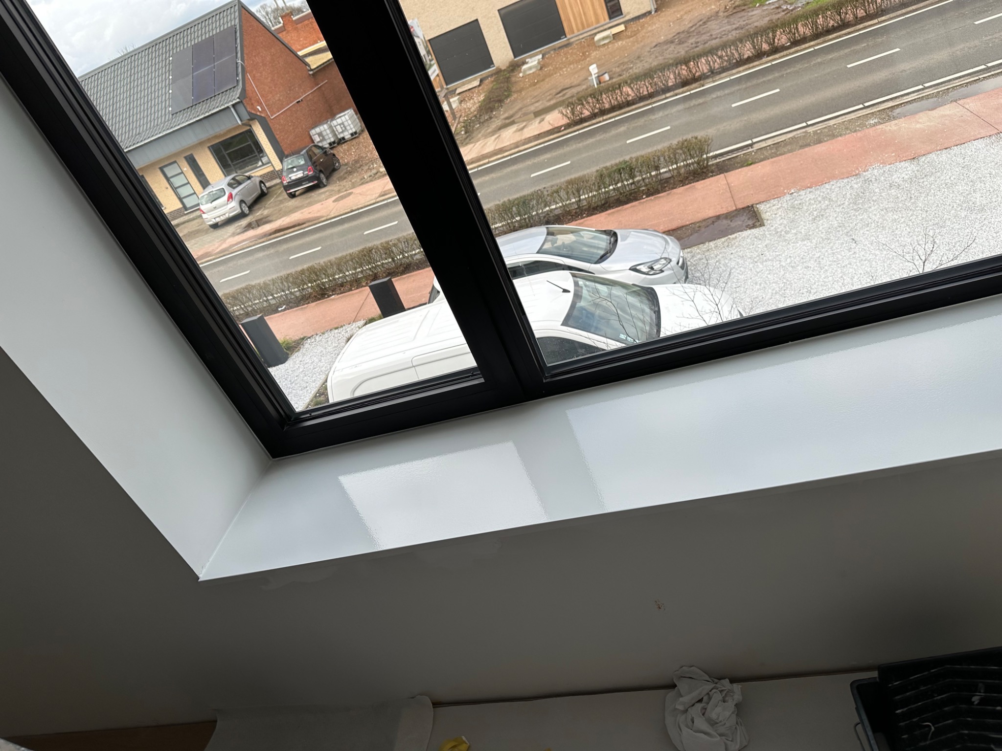 Wnętrze z oknem dachowym z czarną ramą, świeżo pomalowane na biało, widok na ulicę z zaparkowanymi samochodami i budynkami.