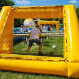 Żółto-niebieska dmuchana arena do gry w piłkarzyki z siatką, wewnątrz której widać grających ludzi i piłkę, otoczona zieloną trawą i drzewami w tle.