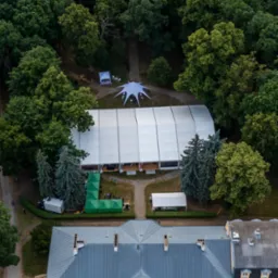 Widok z góry na duży biały namiot imprezowy z zielonymi namiotami cateringowymi, usytuowany obok klasycystycznego pałacu z kolumnami, otoczony zielenią drzew. Dodatkowo widoczny biały namiot...