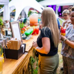 Obsługa barmańska na imprezie plenerowej: bar z drewnianym blatem, kolorowe drinki z owocami, goście z koktajlami, dekoracje roślinne i oświetlenie.