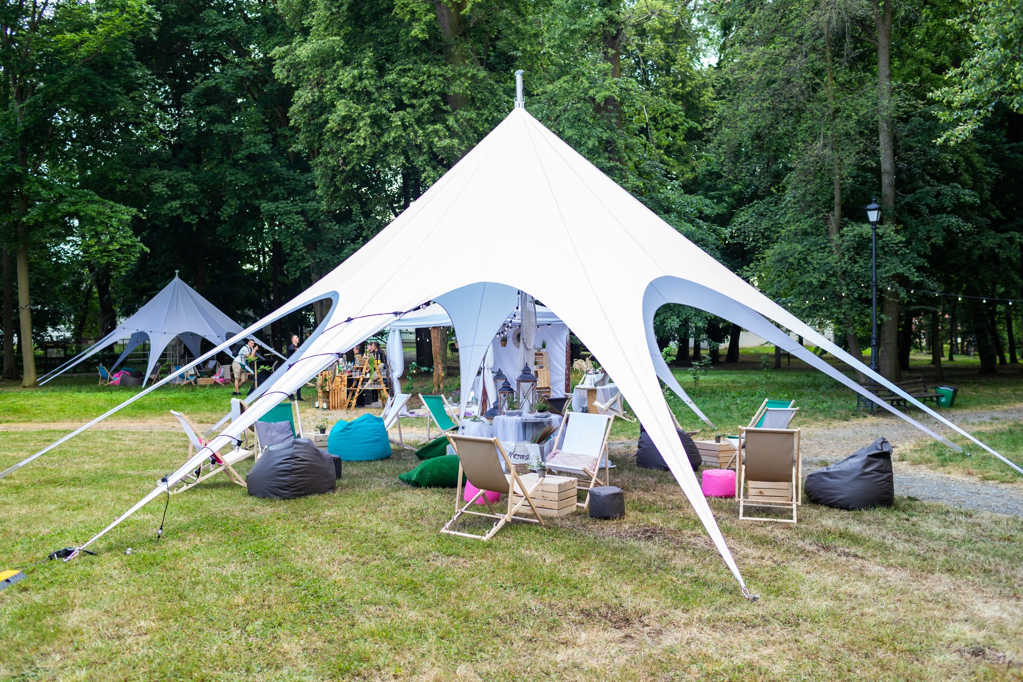 Biały namiot imprezowy rozstawiony na trawie, z widocznymi leżakami, pufami i dekoracjami, tworzący strefę relaksu na świeżym powietrzu, z drugim namiotem w tle.