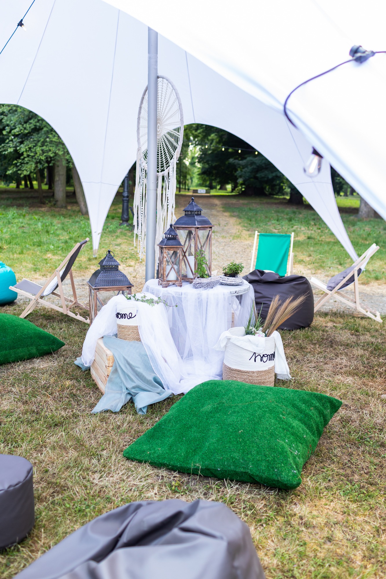 Aranżacja plenerowa pod białym namiotem z lampionami, poduszkami imitującymi trawę, pufami i leżakami tworząca strefę relaksu.