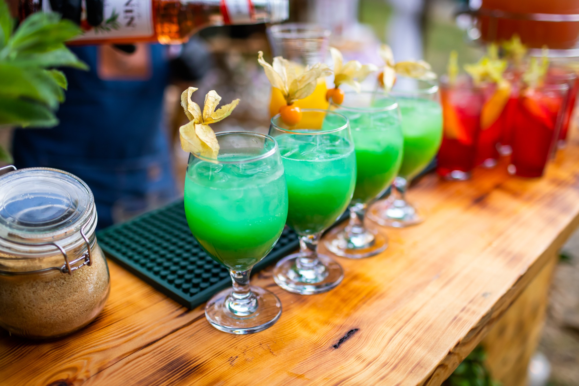 Przygotowywanie kolorowych drinków na drewnianym barze, ozdobionych physalis, z widocznym słoikiem z cukrem trzcinowym i butelką syropu.