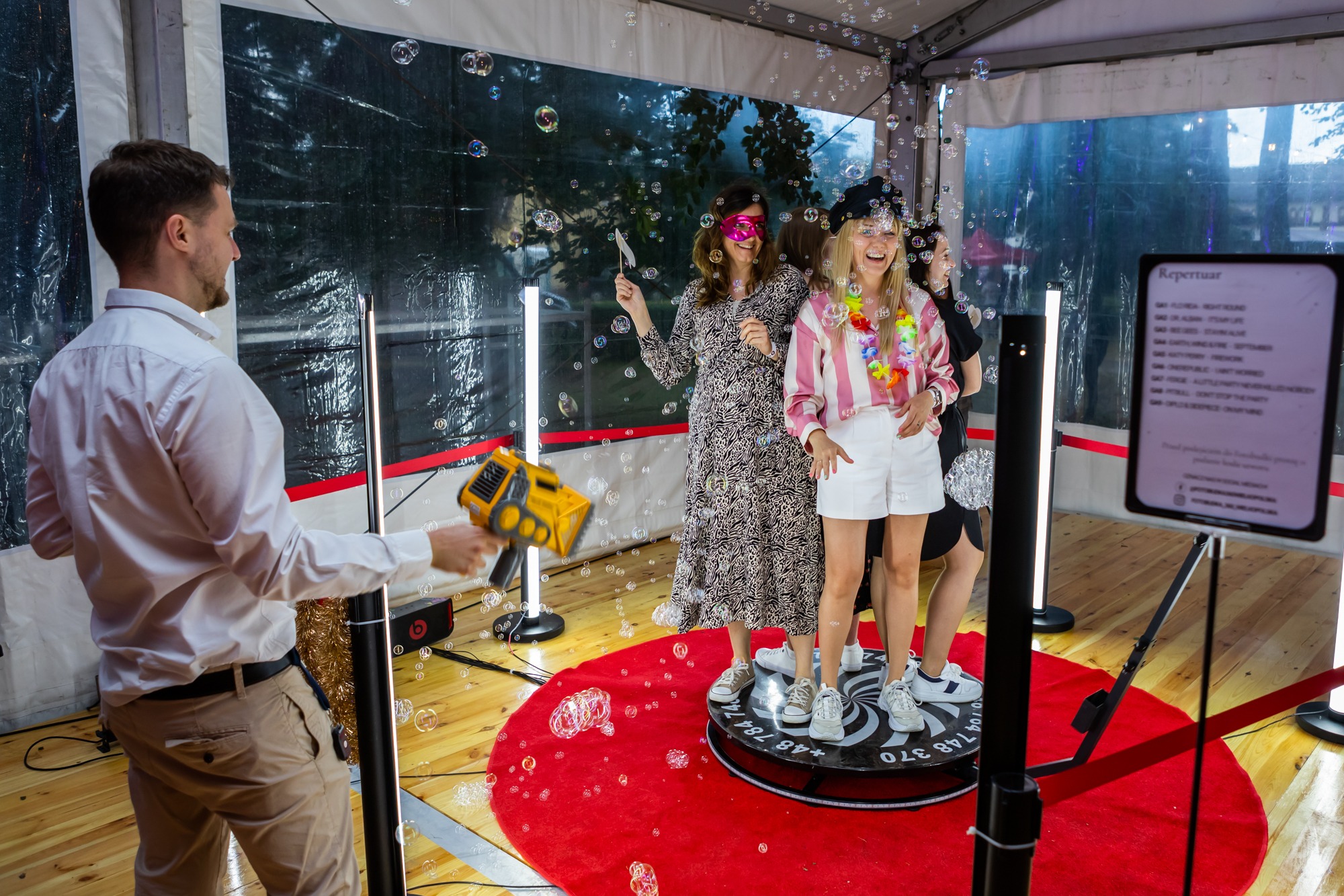 Uśmiechnięte kobiety w maskaradzie pozują na obrotowej platformie podczas imprezy, mężczyzna obsługuje wytwornicę baniek mydlanych, w tle widoczny ekran z repertuarem muzycznym.