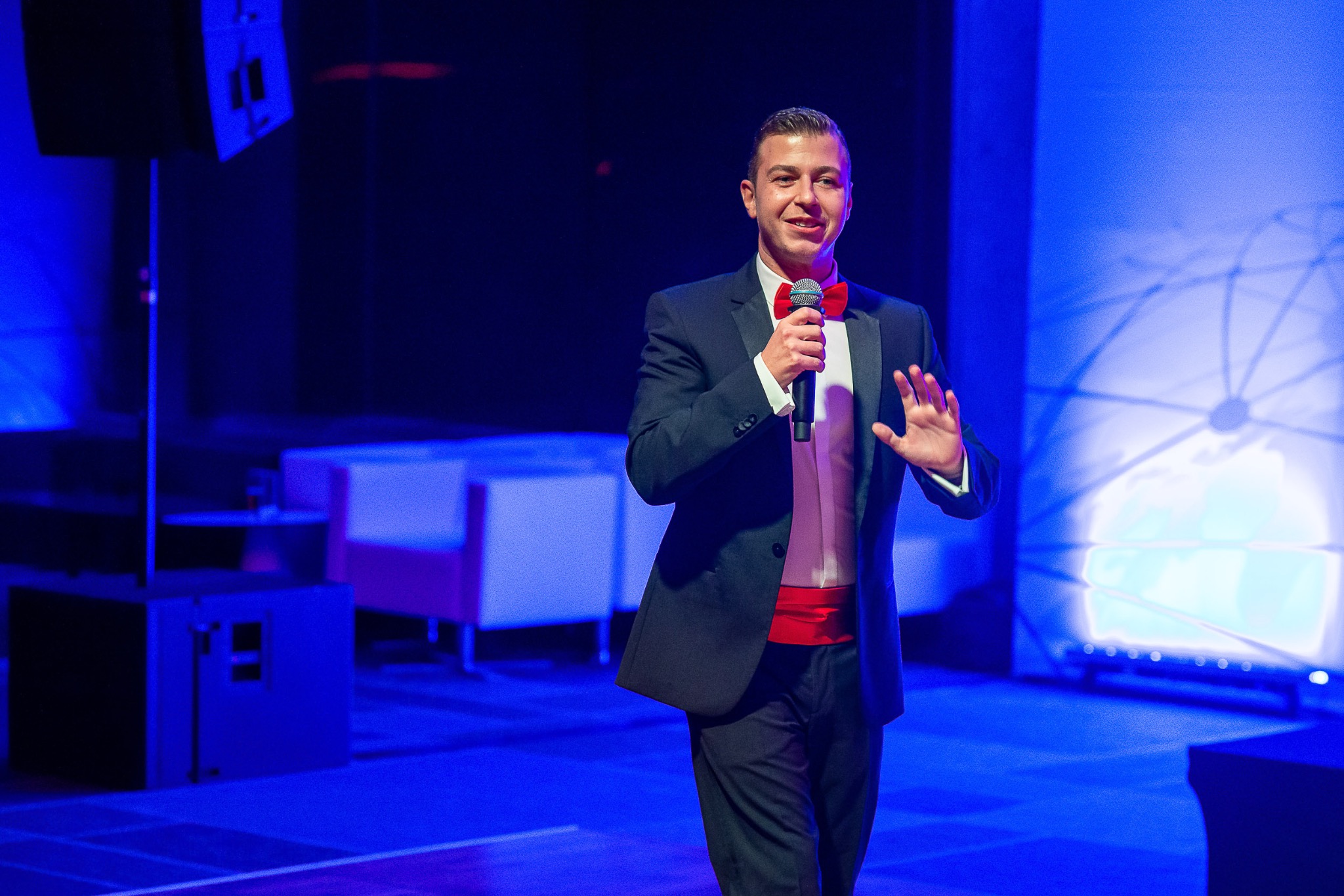 Mężczyzna w smokingu z czerwoną muchą i pasem, trzymający mikrofon i przemawiający na scenie z niebieskim oświetleniem.