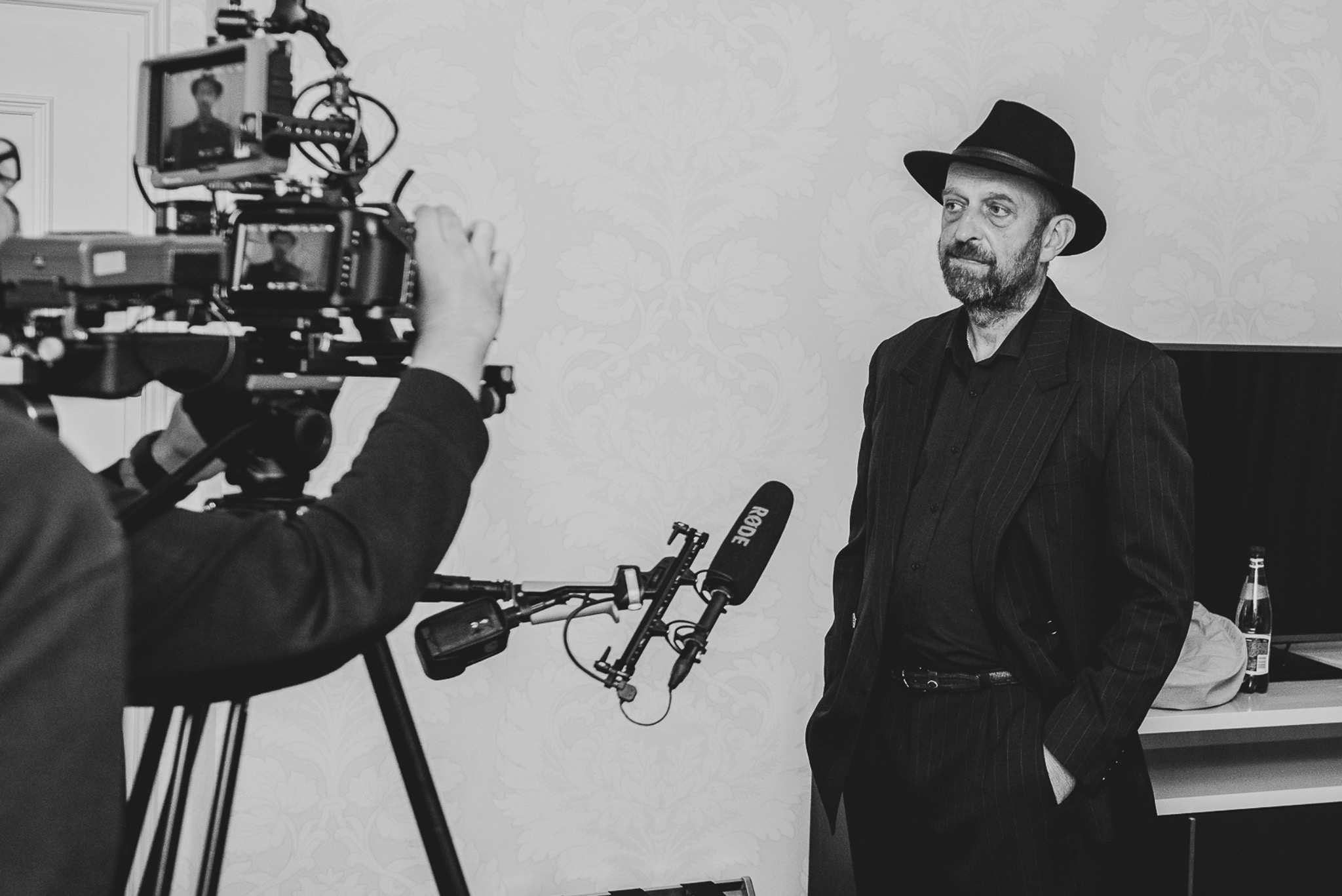 Czarno-białe zdjęcie z planu filmowego: operator kamery z profesjonalnym sprzętem nagrywa mężczyznę w kapeluszu, stojącego na tle ściany z wzorem.