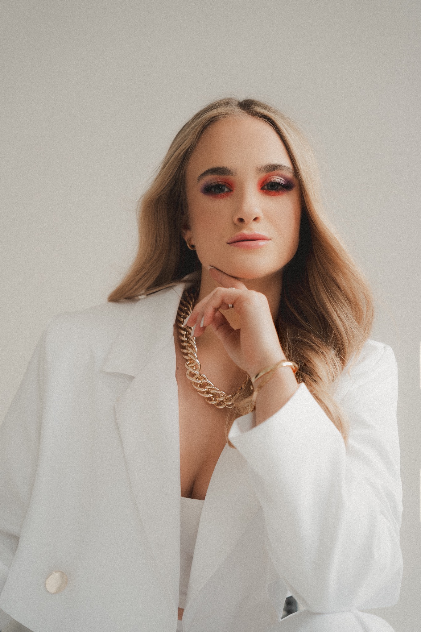 Portret kobiety w białym żakiecie i złotej biżuterii, z wyrazistym makijażem oczu w odcieniach czerwieni i fioletu, pozującej na jasnym tle.