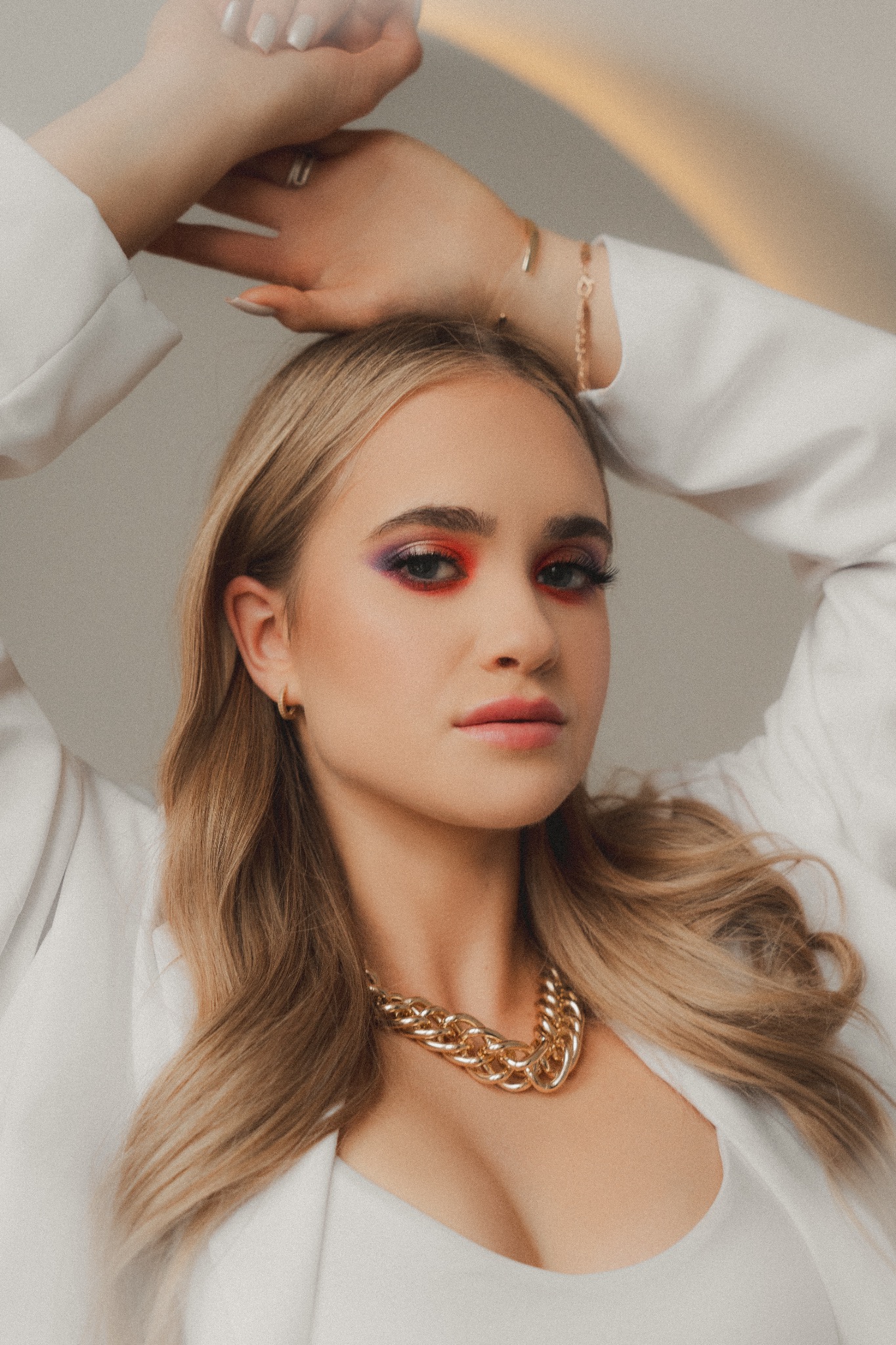 Portret młodej kobiety z wyrazistym makijażem oczu w odcieniach czerwieni i fioletu, ubrana w białą marynarkę i gruby złoty łańcuch, ręce uniesione nad głową, subtelne oświetlenie.