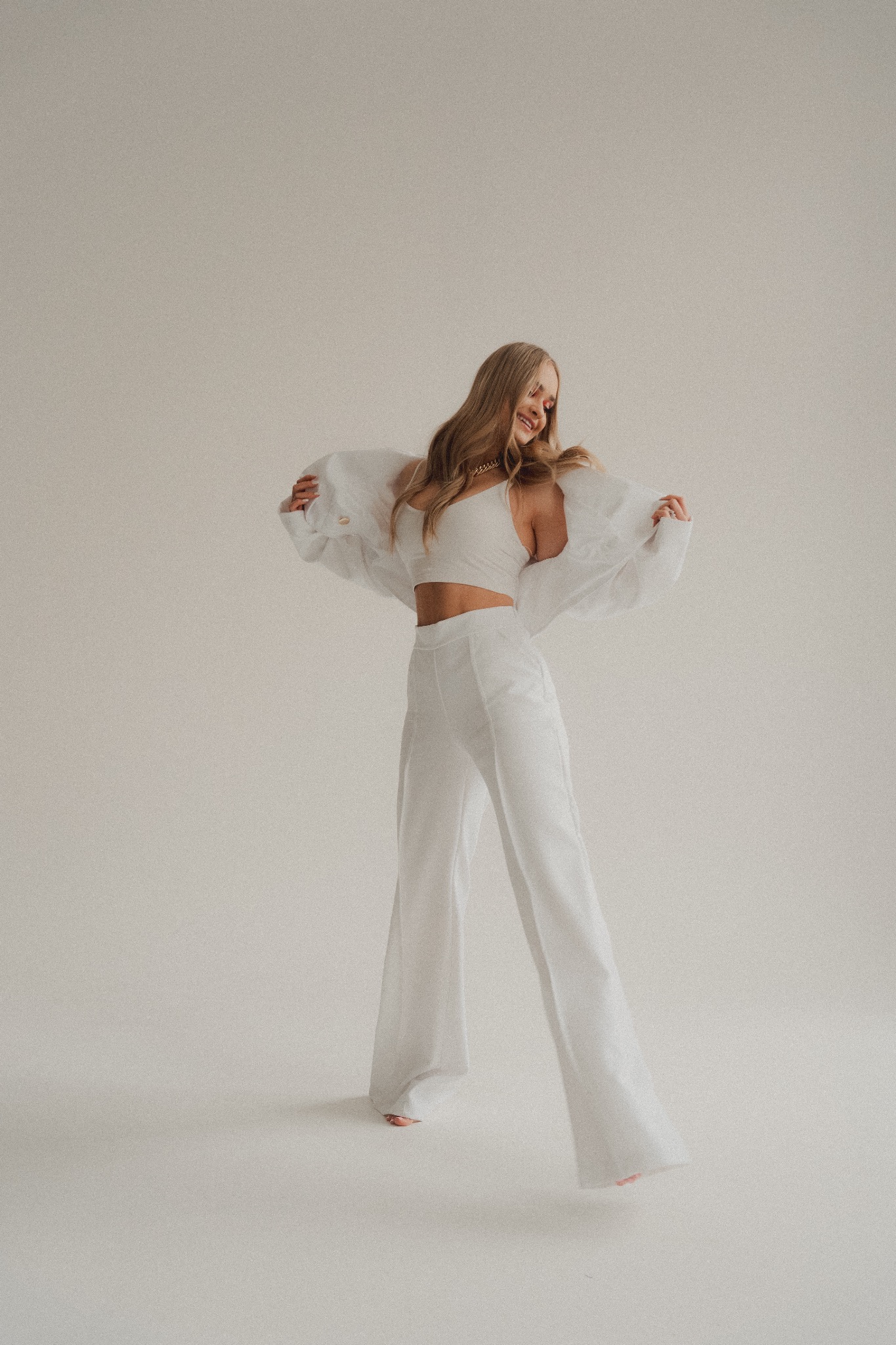 Uśmiechnięta blondynka w białym komplecie (top, spodnie, marynarka) pozuje na jasnym tle, lekko unosząc ręce.