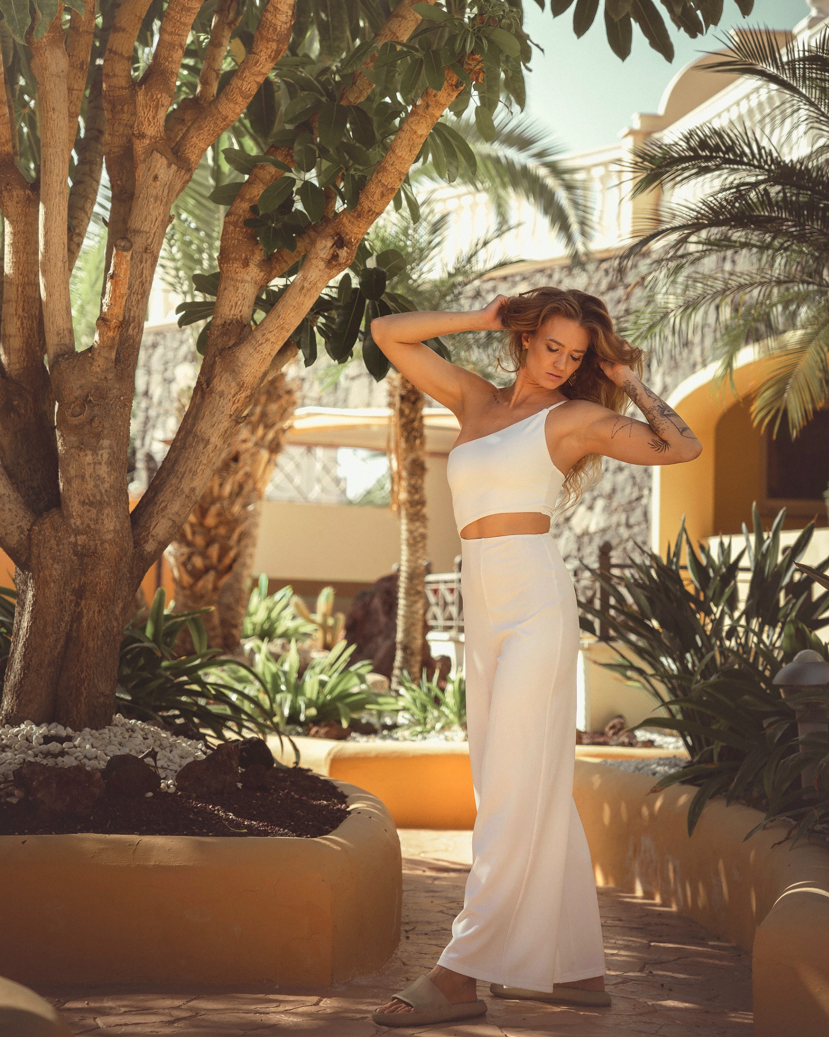 Kobieta w białym kombinezonie na tle egzotycznej roślinności i architektury Teneryfy, dłońmi poprawia włosy, widoczny tatuaż na ramieniu.