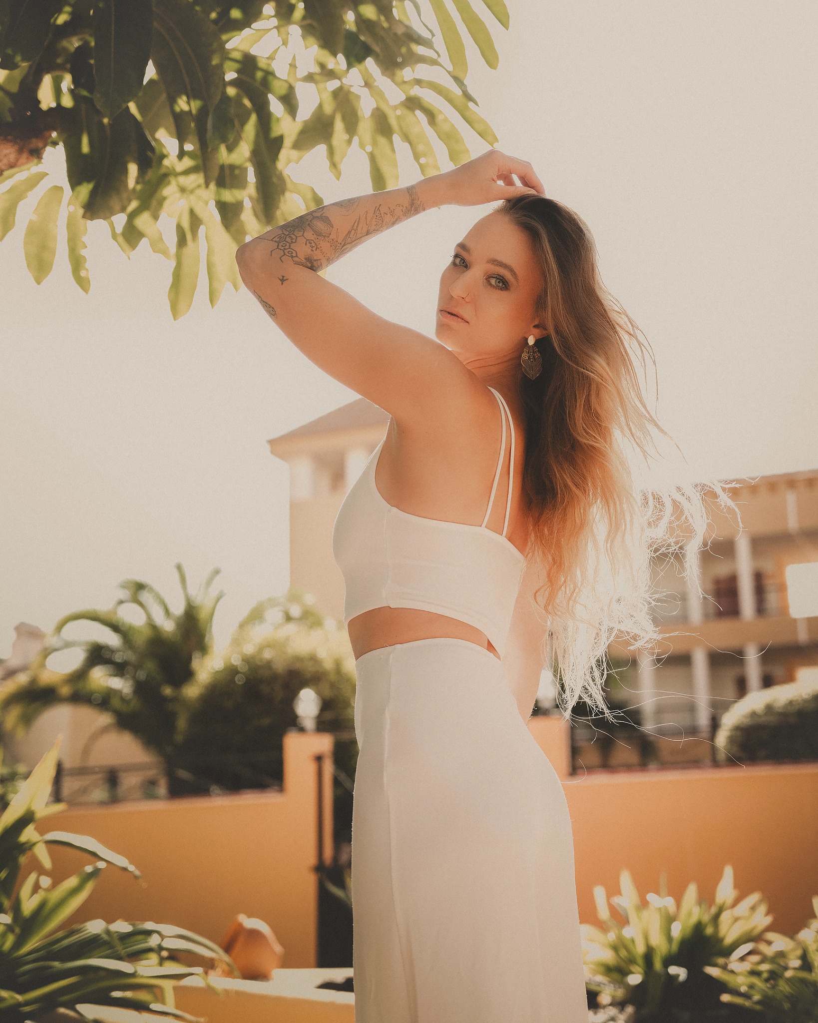 Portret kobiety w białym stroju na tle tropikalnej roślinności i budynku na Teneryfie, z tatuażem na ramieniu i kolczykami w stylu boho.