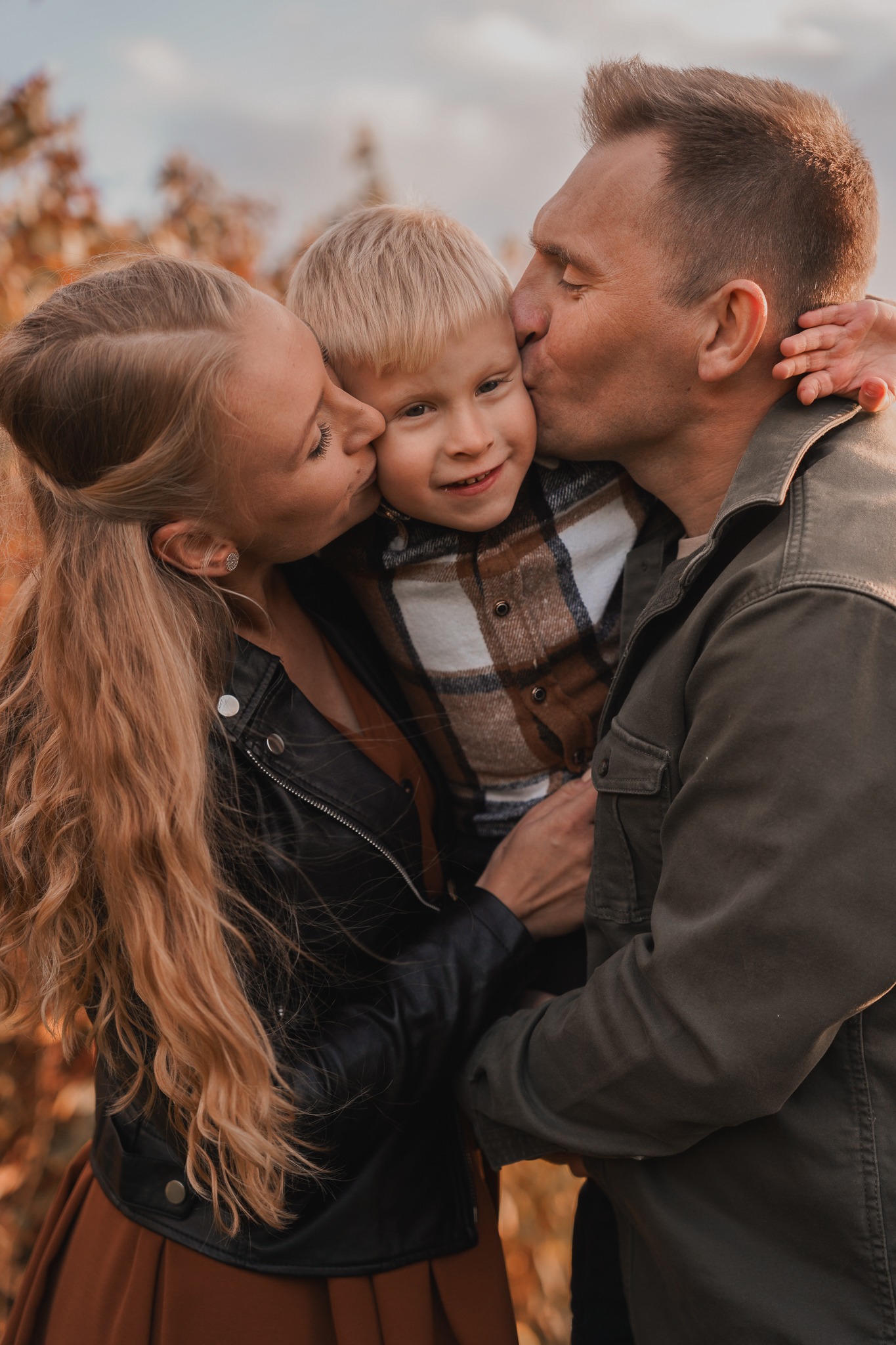 Jesienne ujęcie: mama w czarnej skórzanej kurtce i tata w zielonej kurtce całują w policzki uśmiechniętego blond syna w brązowo-białej koszuli w kratę na tle drzew z brązowymi liśćmi.