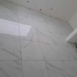 Ściana wyłożona dużymi, białymi płytkami imitującymi marmur, z widocznymi fugami i otworami na instalacje elektryczne w górnej części, pod oknem dachowym.