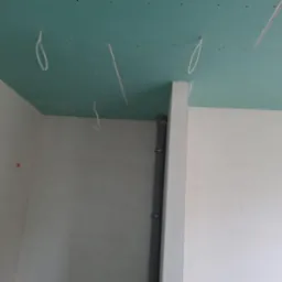 Ściana i sufit pokryte zieloną płytą kartonowo-gipsową, widoczne przewody elektryczne zwisające z sufitu oraz pionowa rura kanalizacyjna przy ścianie.