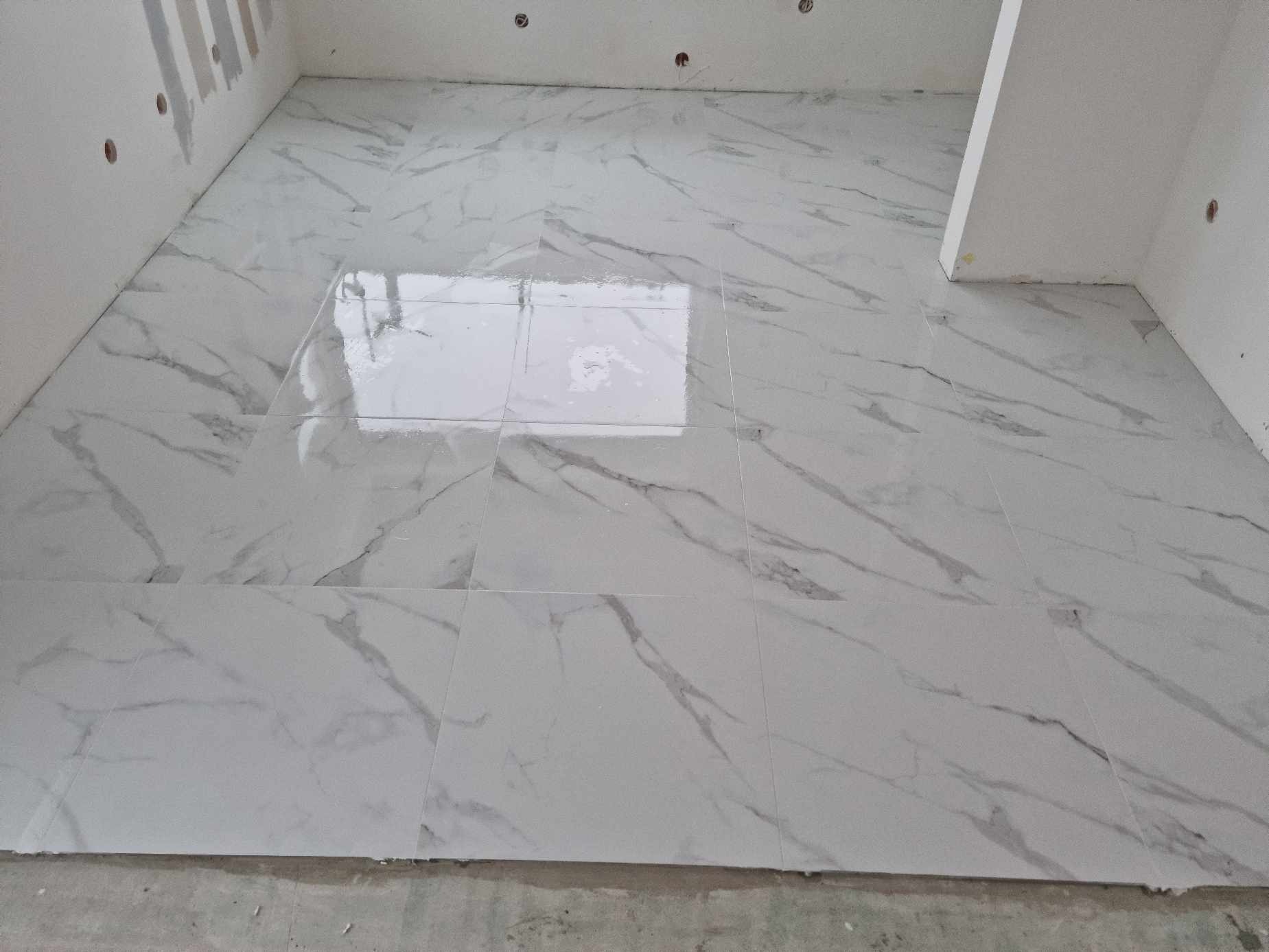 Świeżo ułożone, błyszczące, białe płytki podłogowe z szarym marmurkowym wzorem, częściowo mokre po myciu, na tle surowych ścian z widocznymi punktami elektrycznymi.