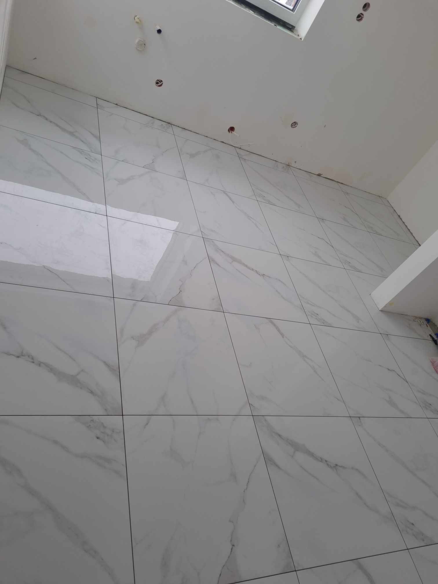 Ściana wyłożona dużymi, białymi płytkami imitującymi marmur, z widocznymi fugami i otworami na instalacje elektryczne w górnej części, pod oknem dachowym.