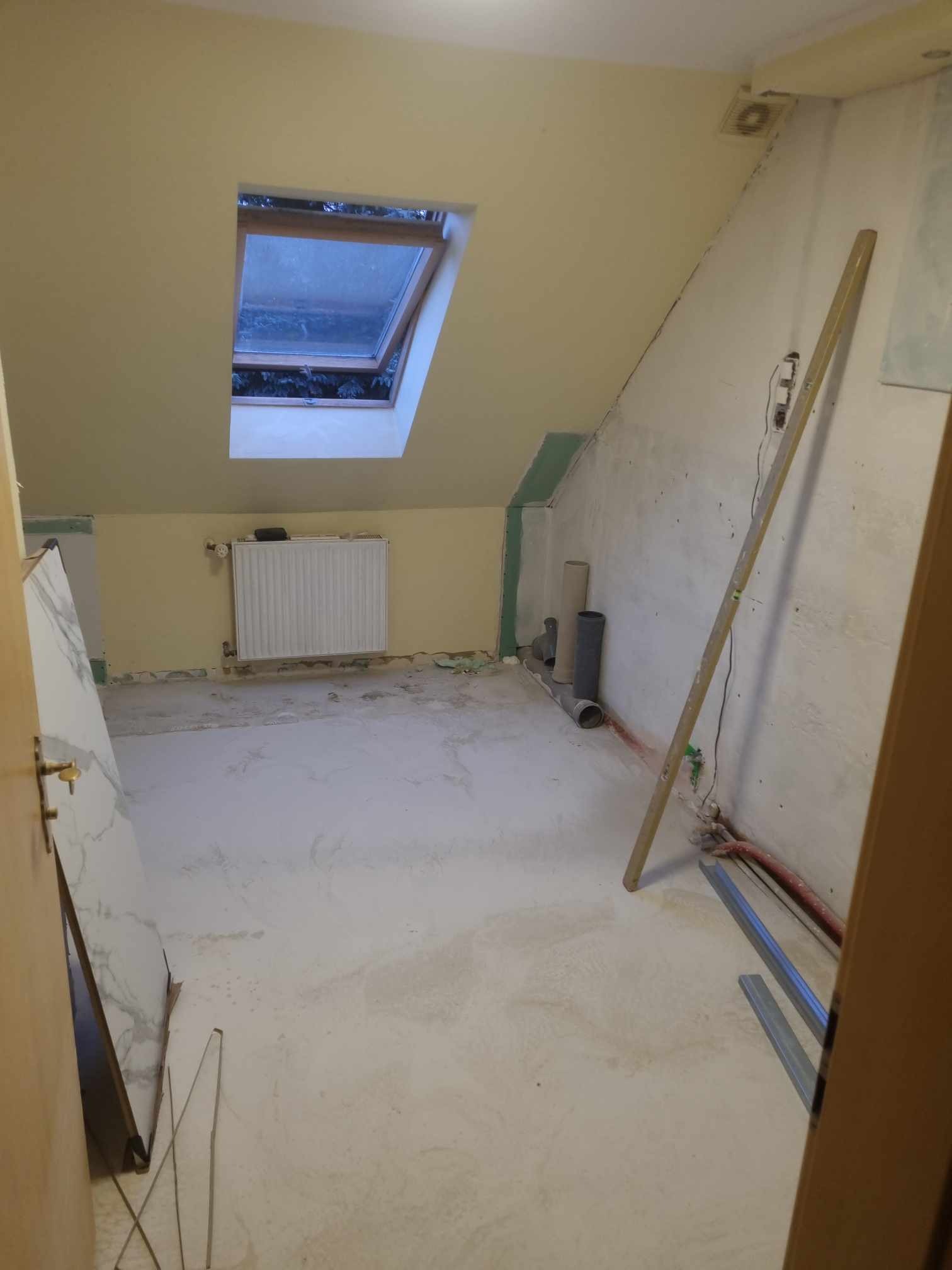 Pokój na poddaszu w trakcie remontu: widoczne rury, płyty karton gips, grzejnik, okno dachowe i poziomica oparta o ścianę.