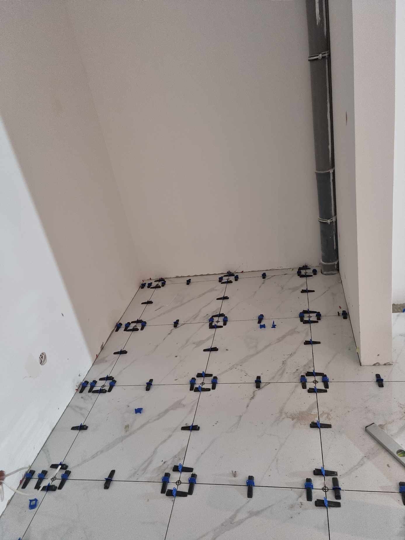 Układanie płytek podłogowych z użyciem systemu poziomowania; widoczne krzyżyki dystansowe, kliny i poziomica; w tle rura kanalizacyjna przy ścianie.