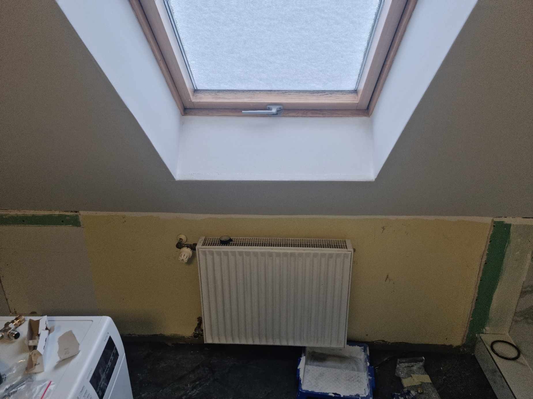 Remont łazienki na poddaszu: widok na okno dachowe z widocznym śniegiem, grzejnik, fragment ściany z żółtą farbą i przygotowane narzędzia malarskie.