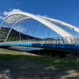 INGVARR SPÓLKA Z O O - Łukowy most dla pieszych z białą konstrukcją i niebieskim spodem, z metalowymi barierkami, na tle zielonych wzgórz i błękitnego nieba z chmurami.