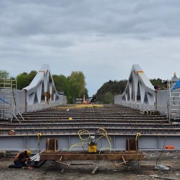 INGVARR SPÓLKA Z O O - Spawacz w pomarańczowej kamizelce i żółtym kasku spawa element konstrukcji mostu na tle szarego nieba i drzew