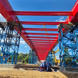 INGVARR SPÓLKA Z O O - Konstrukcja stalowa mostu kolejowego w trakcie budowy, pomalowana na czerwono i niebiesko, widok z dołu na tle błękitnego nieba.