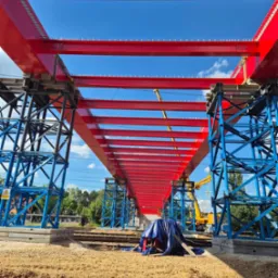 Konstrukcja stalowa mostu kolejowego w trakcie budowy, pomalowana na czerwono i niebiesko, widok z dołu na tle błękitnego nieba.