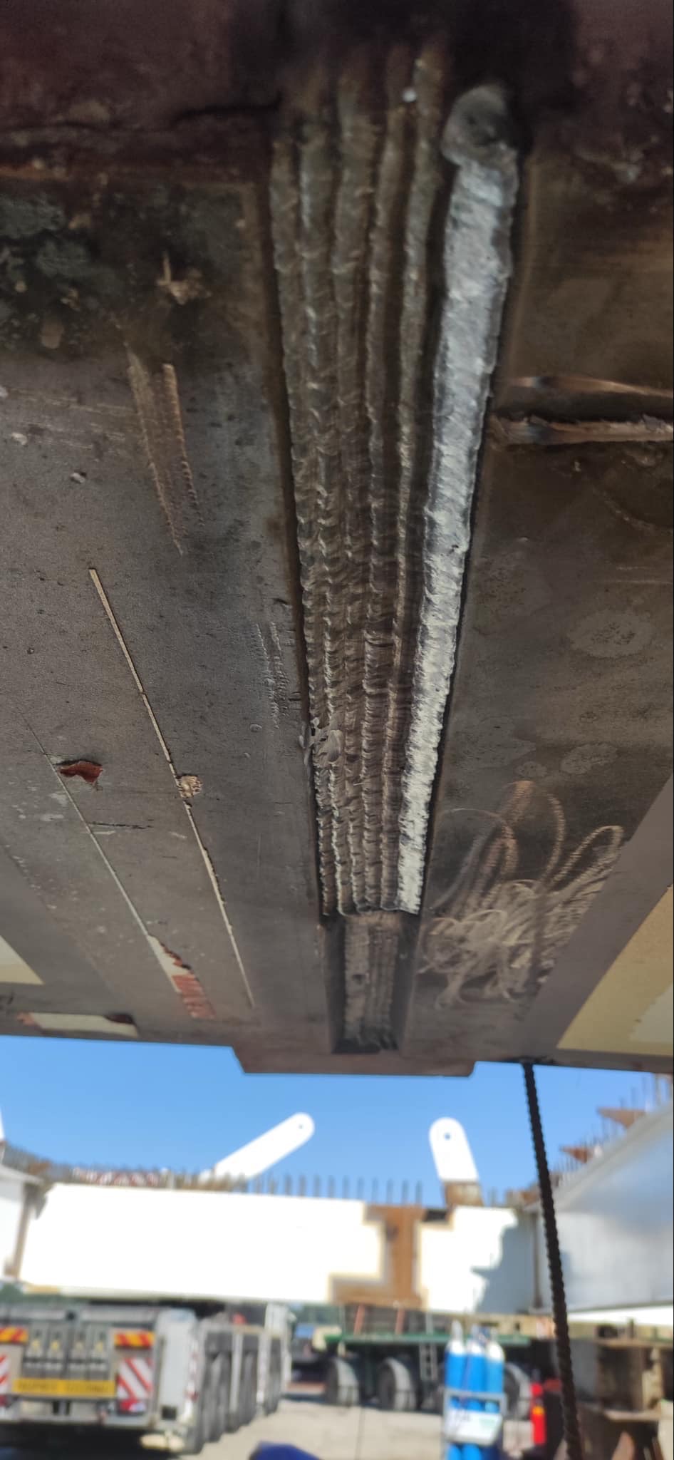 Spoina wielościegowa na metalowej konstrukcji, widoczne ślady po spawaniu oraz fragment transportu konstrukcji w tle.