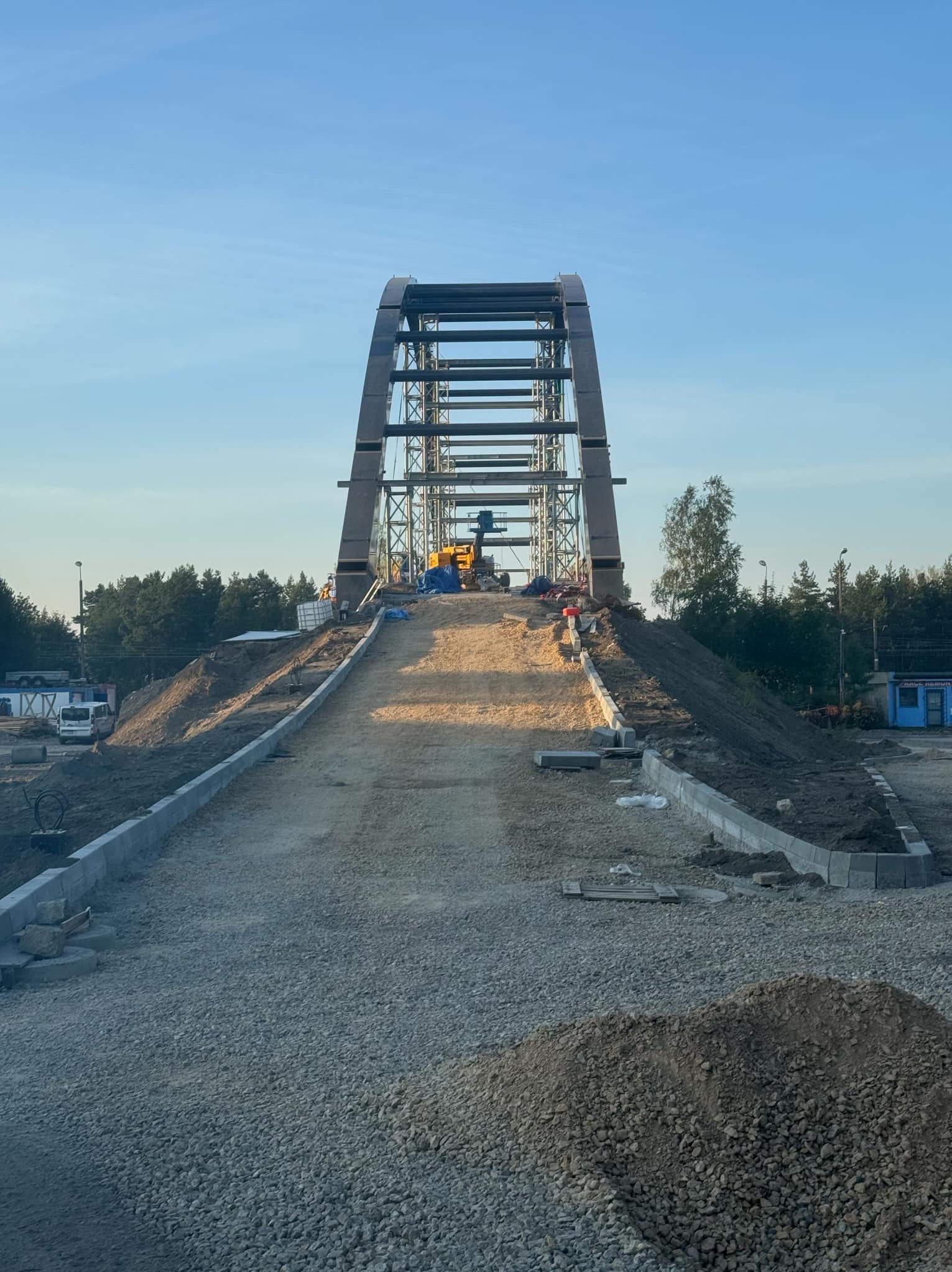 Konstrukcja metalowego mostu łukowego w trakcie budowy, widoczny sprzęt budowlany i przygotowane podłoże z kamienia