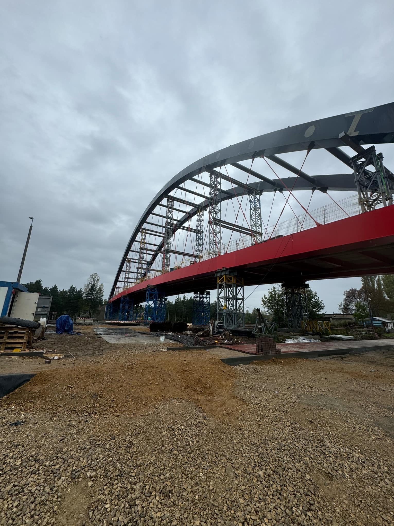 Konstrukcja łukowego mostu stalowego w trakcie budowy, widoczne rusztowania i elementy konstrukcyjne pomalowane na czerwono i niebiesko, teren budowy z gruzem i piaskiem, pochmurne niebo.