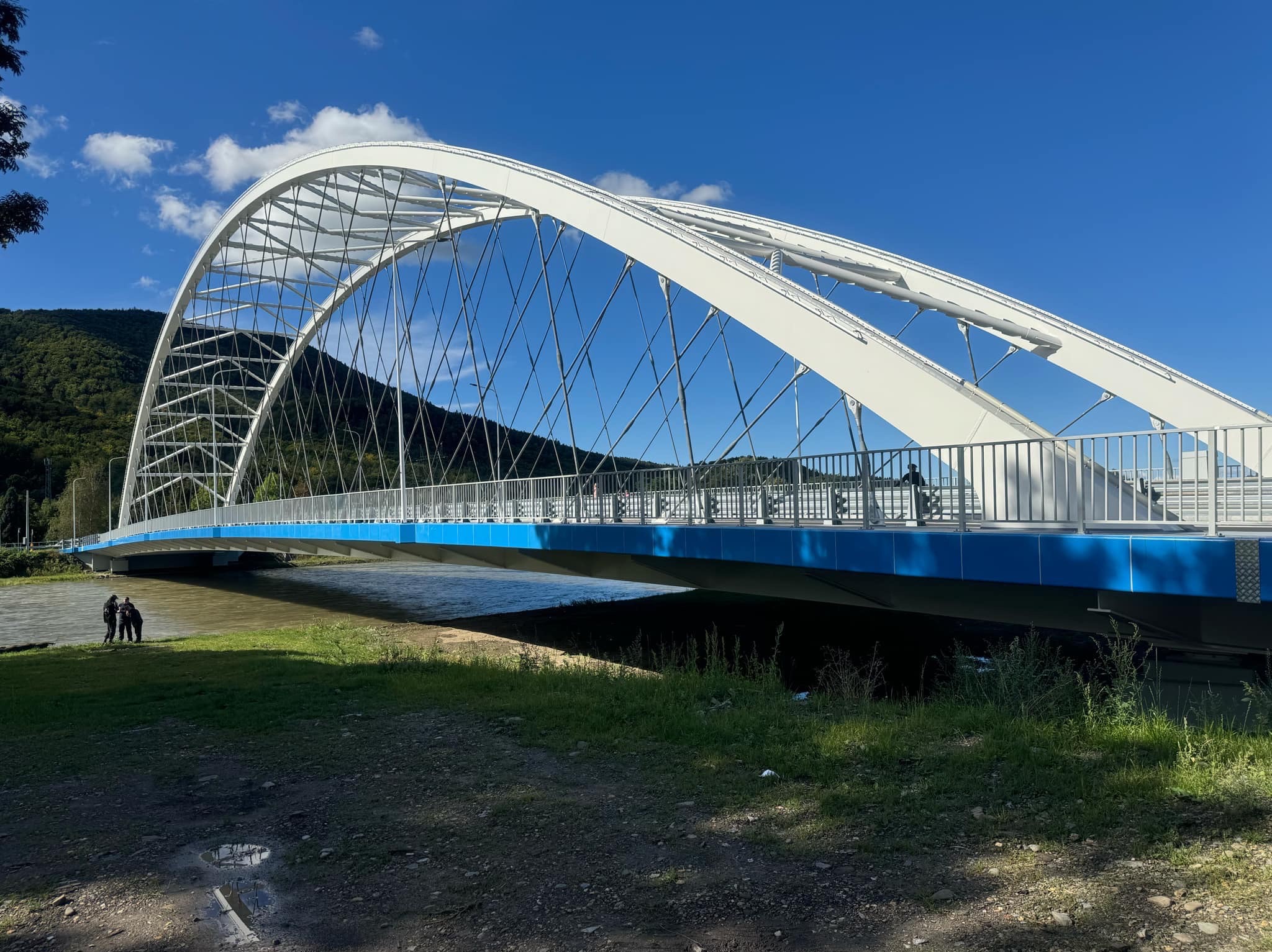 Łukowy most dla pieszych z białą konstrukcją i niebieskim spodem, z metalowymi barierkami, na tle zielonych wzgórz i błękitnego nieba z chmurami.