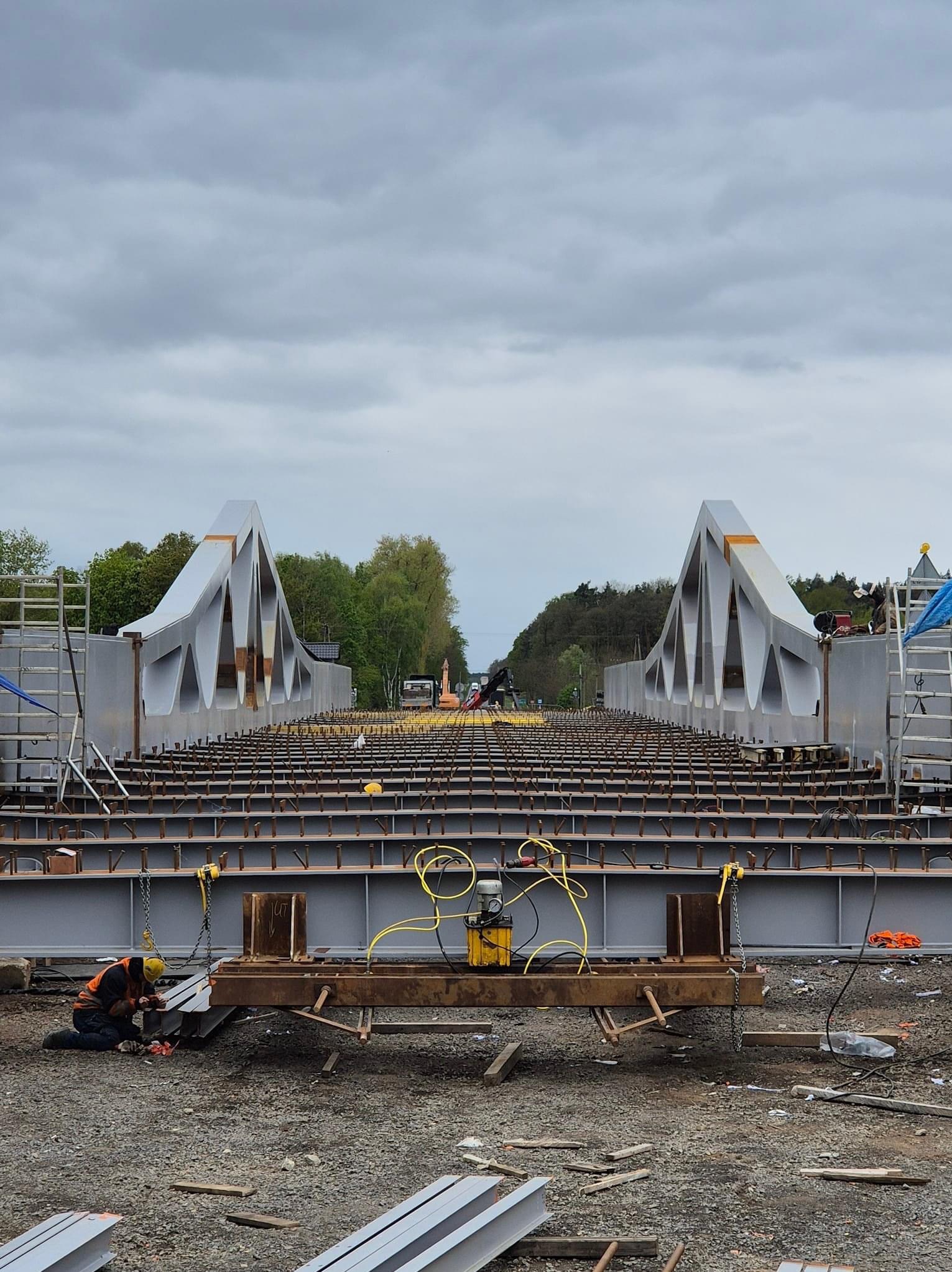 Spawacz w pomarańczowej kamizelce i żółtym kasku spawa element konstrukcji mostu na tle szarego nieba i drzew