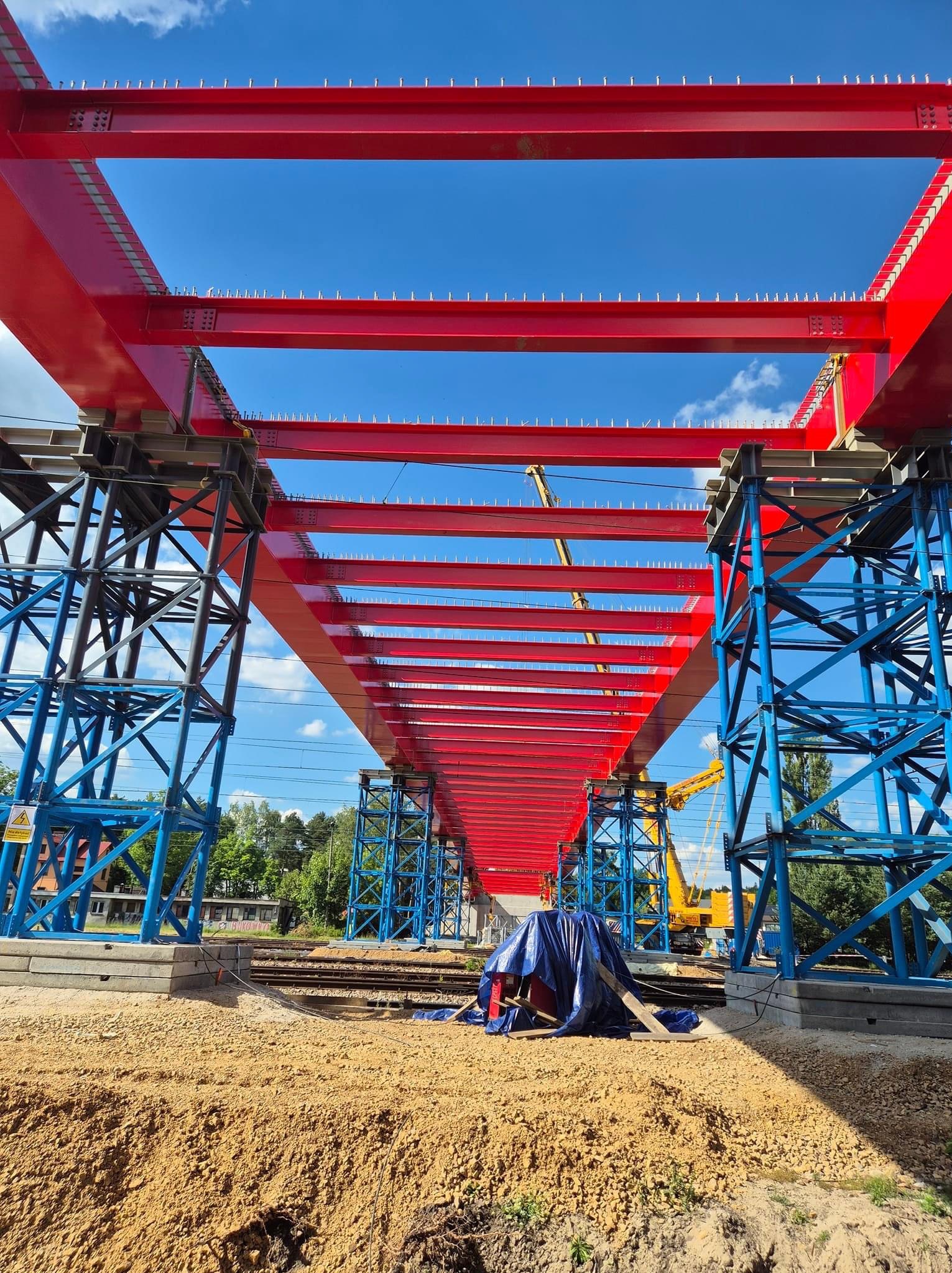 Konstrukcja stalowa mostu kolejowego w trakcie budowy, pomalowana na czerwono i niebiesko, widok z dołu na tle błękitnego nieba.