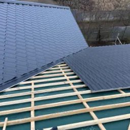 Montaż grafitowej blachodachówki na drewnianej konstrukcji dachu, widoczni dekarze w trakcie pracy, zabezpieczenie w postaci membrany dachowej.