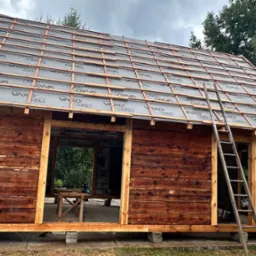 Budowa drewnianej konstrukcji z widoczną więźbą dachową pokrytą membraną izolacyjną, drabina oparta o budynek, teren budowy z drzewami w tle i pochmurnym niebem.