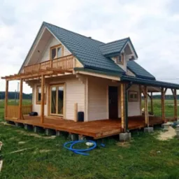Nowoczesny, drewniany dom z ciemnozielonym dachem z blachy trapezowej, z drewnianym tarasem i balkonem, posadowiony na betonowych bloczkach, na zewnątrz widoczny zwinięty niebieski wąż ogrodowy.