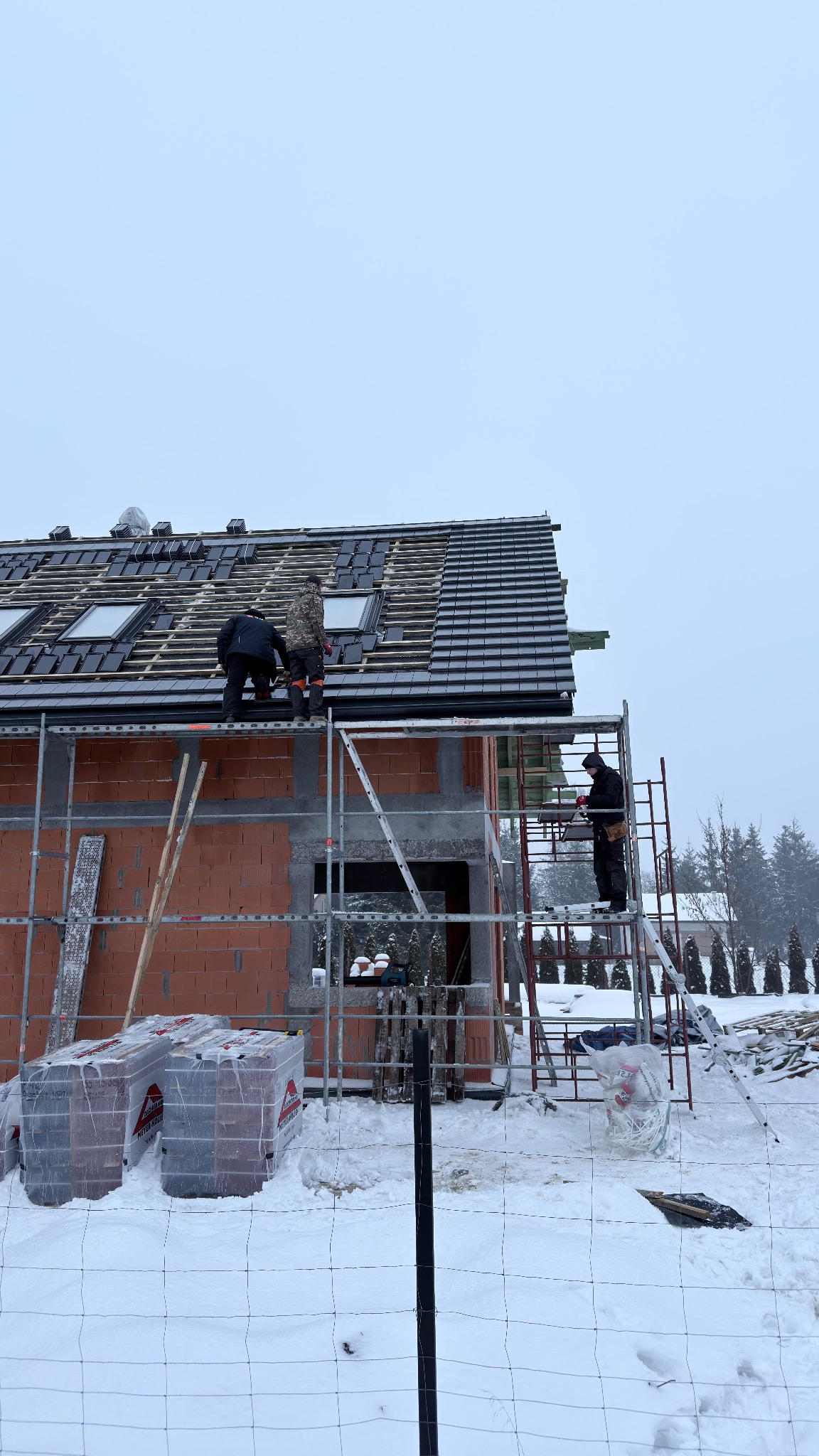 Montaż dachówki na nowym domu w zimowej scenerii. Murarze na rusztowaniu układają dachówkę, widoczne okna dachowe i materiały budowlane. Praca w toku, zimowy krajobraz.