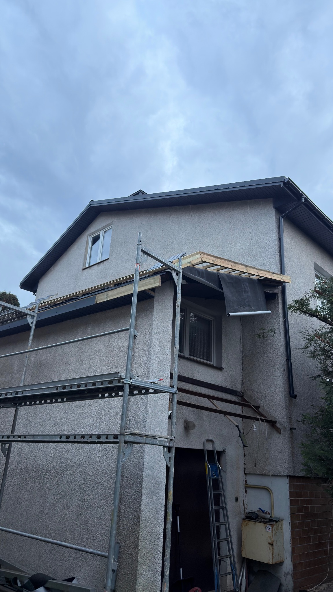 Dom w trakcie budowy z rusztowaniem i częściowo zadaszonym wejściem. Widoczne elementy konstrukcyjne i materiały budowlane.