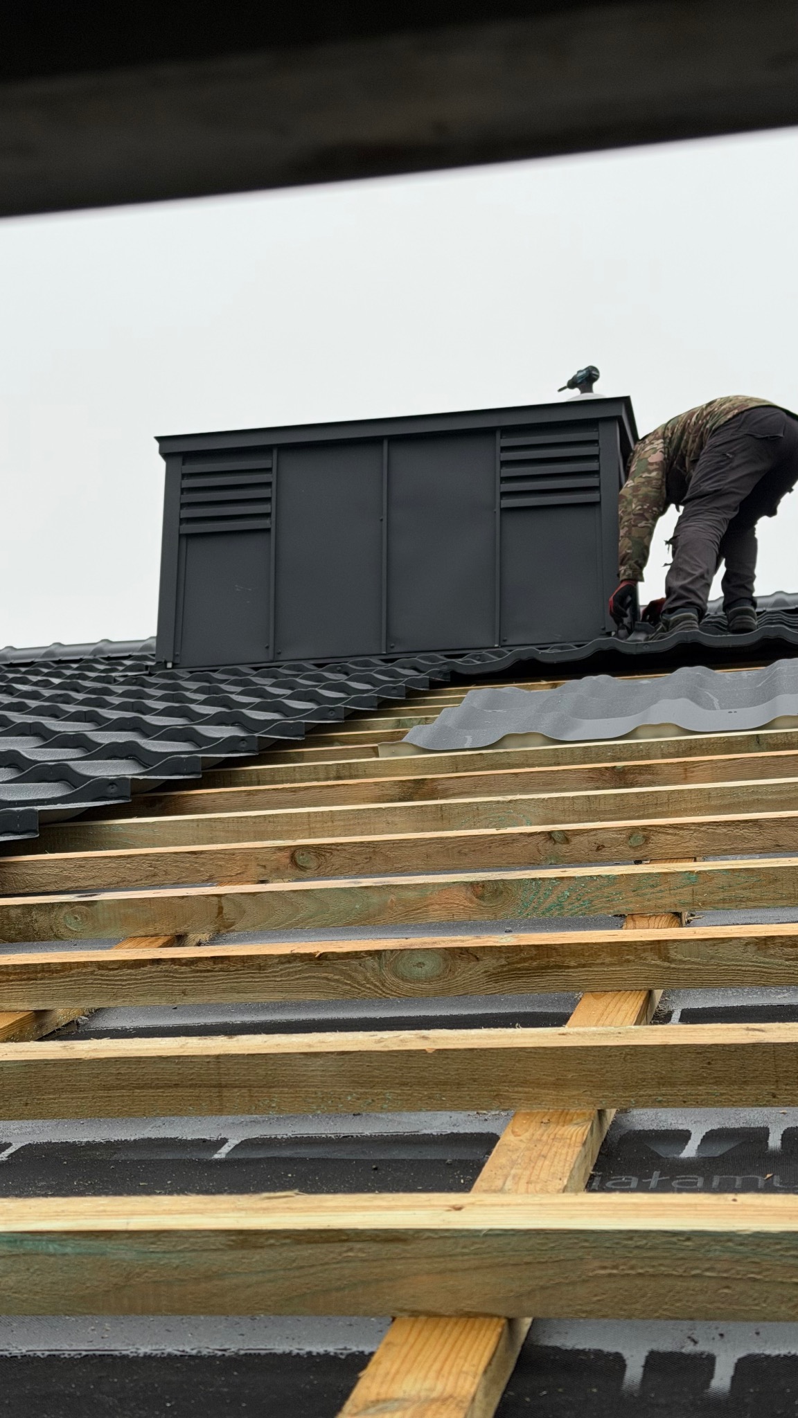 Montaż dachu z czarnej dachówki falistej, widoczna więźba i komin z wentylacją. Dekarz w trakcie pracy, na kominie siedzi ptak. Ujęcie z dołu.