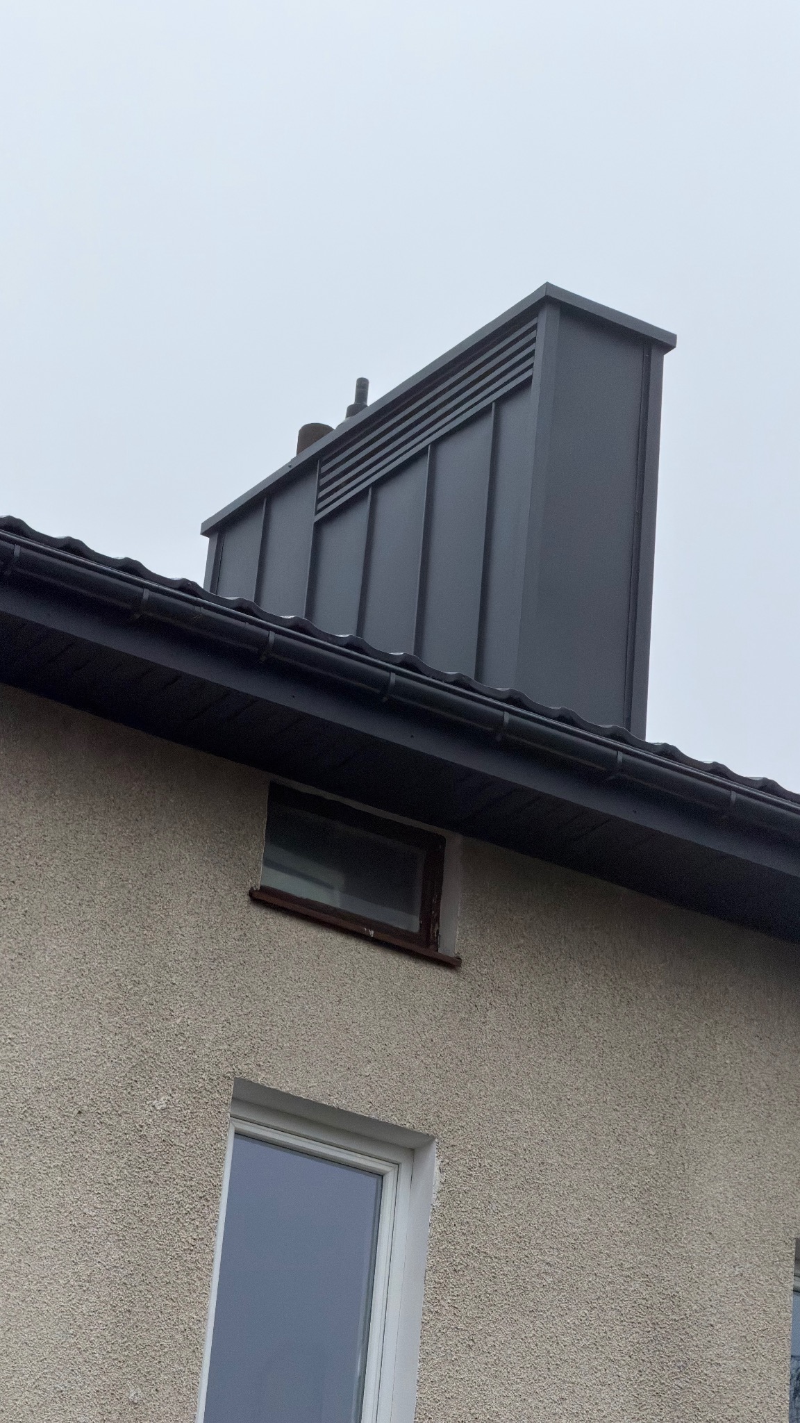 Szary komin wentylacyjny z blachy na tle pochmurnego nieba, widoczny z dołu, z fragmentem dachu i elewacji budynku. Nowoczesne wykończenie.