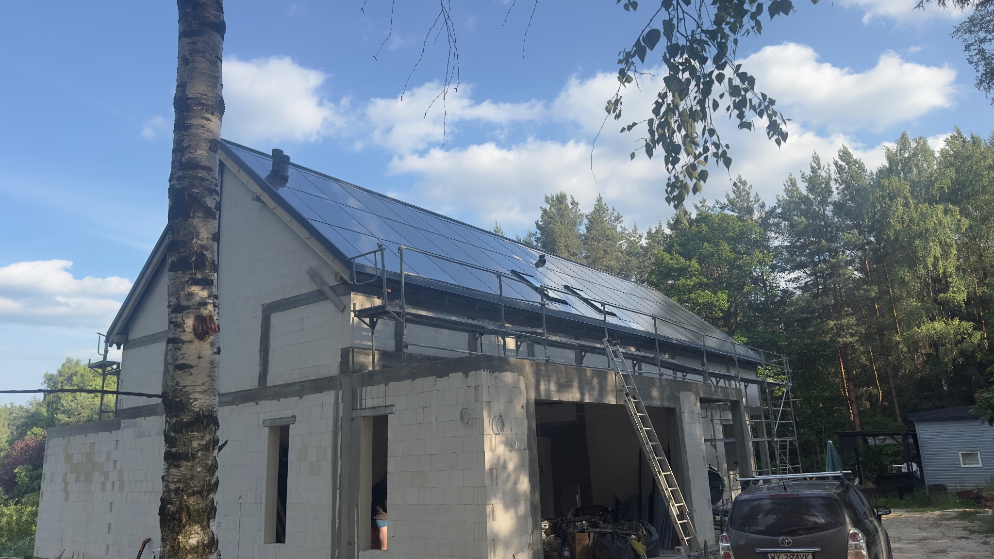 Dom w trakcie budowy z zamontowanymi panelami fotowoltaicznymi na dachu, widoczne rusztowania i drzewo brzozy na pierwszym planie.