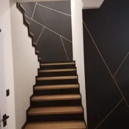 Drewniane schody z czarnymi podstopnicami i geometrycznym wzorem z cienkich białych linii na ciemnoszarej ścianie.