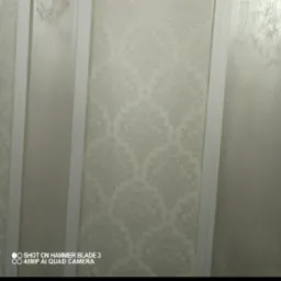 Ściana z dekoracyjną tapetą w odcieniach beżu i szarości, obok fragment gładkiej, pomalowanej ściany oddzielonej białym narożnikiem.